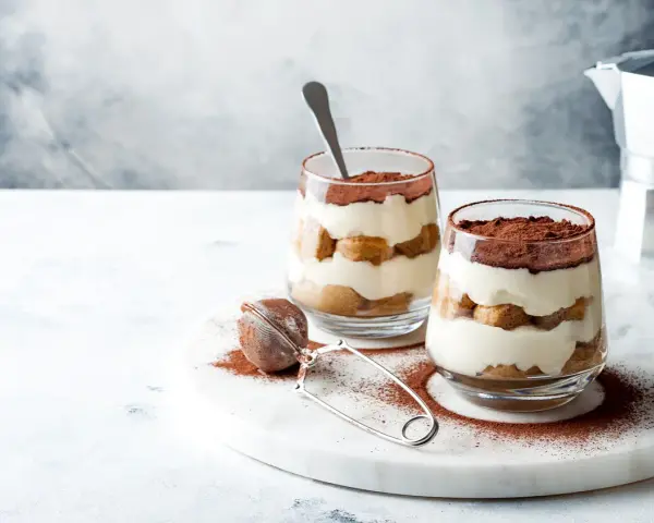 Dwa pucharki z kremowym tiramisu bez jajek, posypane kakao. Obok sitko do kakao i kawiarka.