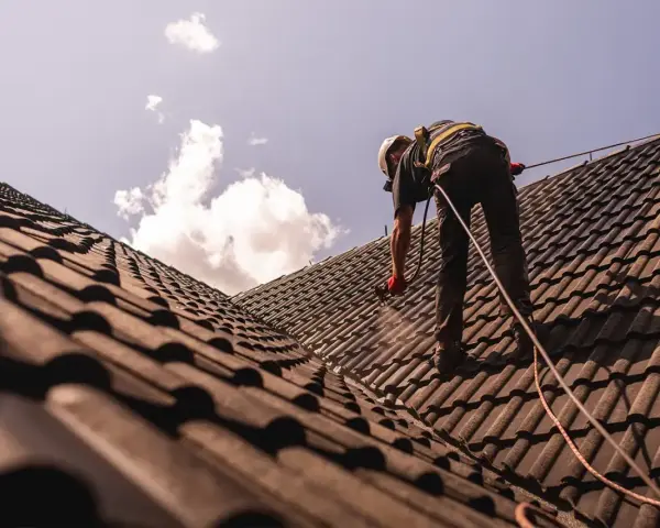Pracownik w kasku i uprzęży zabezpieczającej wykonuje renowację dachu, natryskując dachówkę.