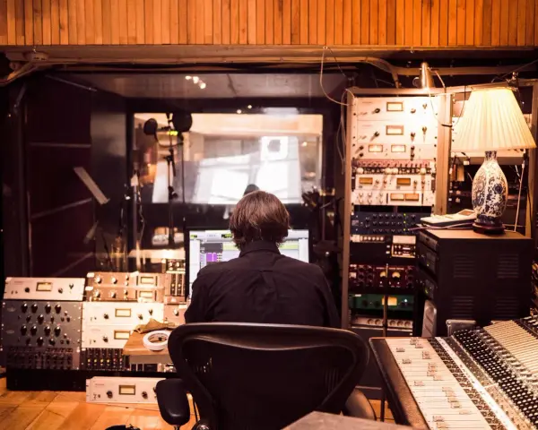 Osoba siedzi przed komputerem w profesjonalnym **recording studio**, otoczona sprzętem audio i mikserem.