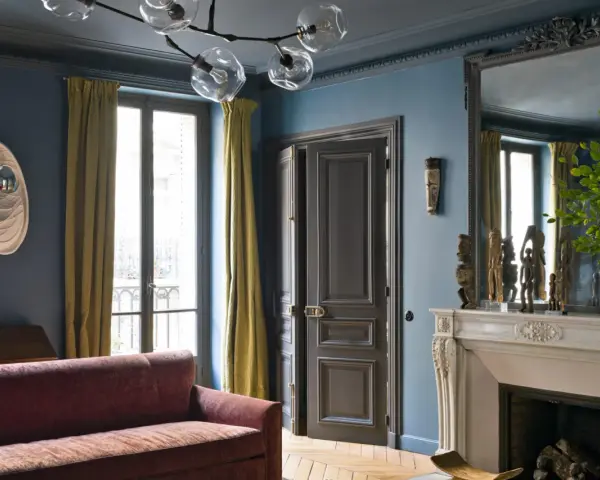 Soggiorno elegante con divano bordeaux, pareti blu balena e tendaggi dorati. Un camino in marmo e sculture africane completano l'arredo.