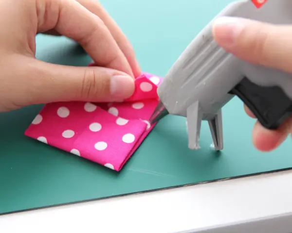 Mani che piegano un tessuto rosa a pois bianchi mentre una pistola per colla a caldo deposita un punto. La colla a caldo si stacca con l'acqua, un dettaglio importante per i lavoretti.