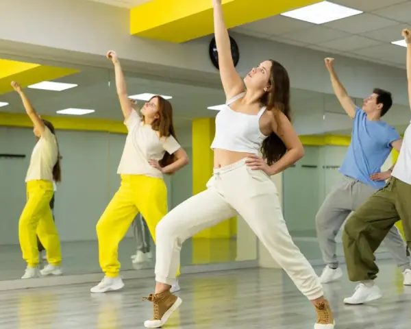 Grupa młodych ludzi ćwiczy taniec w sali z lustrami. Ich ruchy oddają energię gatunku muzycznego pop.