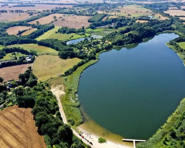 Malownicze jezioro Marszewo otoczone zielenią i polami. Widać piaszczystą plażę z pomostem i kilka samochodów.