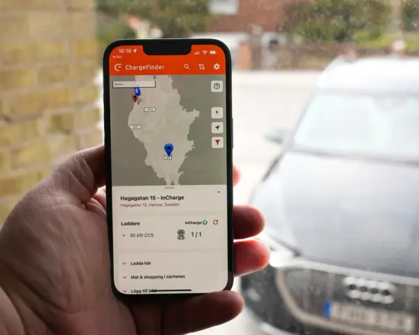 En hand håller upp en mobil med appen ChargeFinder som visar en karta över laddstationer för elbil. En laddapp visar en laddare nära en elbil.