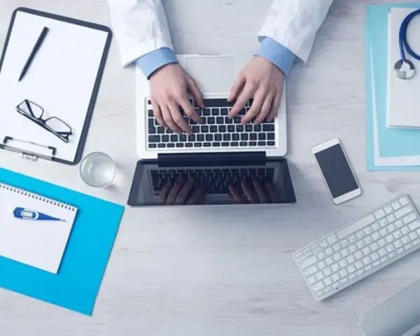 Lekarz pisze na laptopie, obok stetoskop, recepta, telefon i termometr.