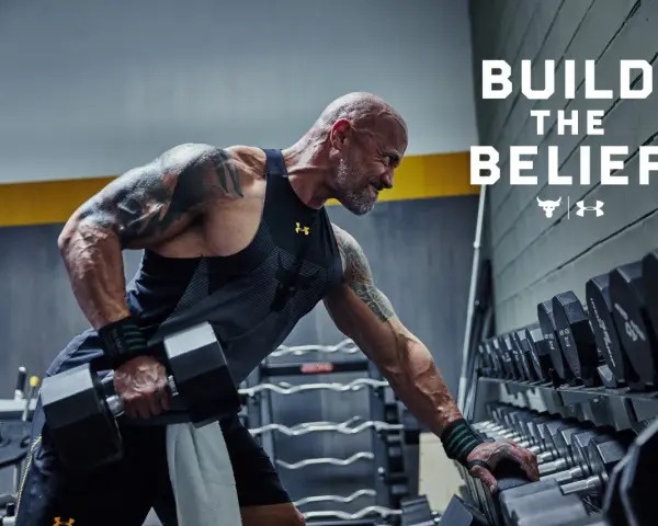 Dwayne "The Rock" Johnson w koszulce Under Armour wykonuje ćwiczenia z hantlami. W tle stojaki z ciężarami.