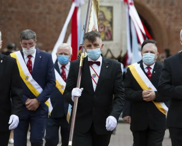 Rycerze Kolumba w garniturach i maskach maszerują z flagą.