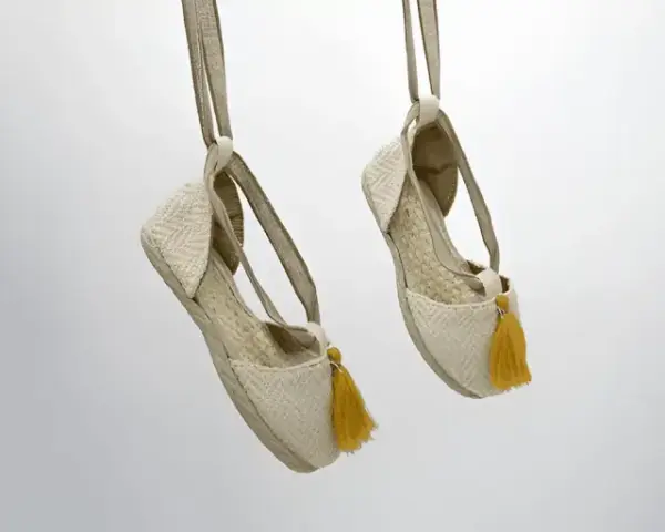 Dos alpargatas de tela beige con pompones amarillos cuelgan de tiras, mostrando su diseño veraniego.