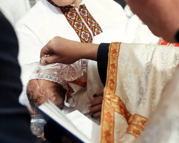 Ksiądz w bogato zdobionej szacie chrzci dziecko, polewając mu głowę wodą z kryształowej karafki.
