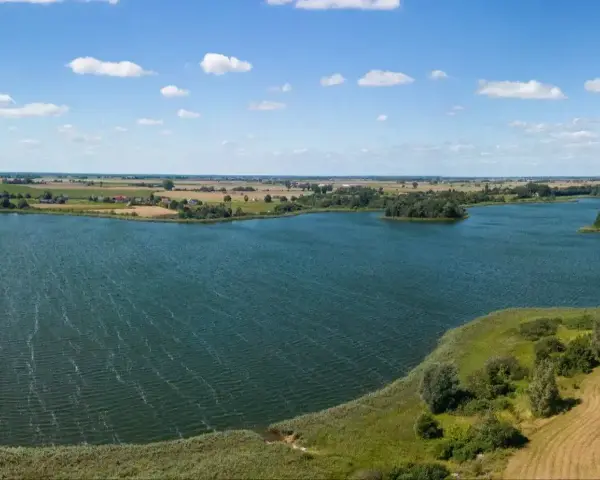 Malownicze jezioro Lednica z wyspą pośrodku, otoczone zielenią i polami. Błękitne niebo z białymi chmurami.