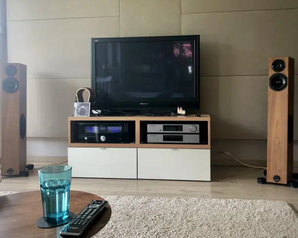 Dwie kolumny Audio Physic Virgo w drewnianej okleinie, telewizor, sprzęt audio i szklanka wody na stoliku.