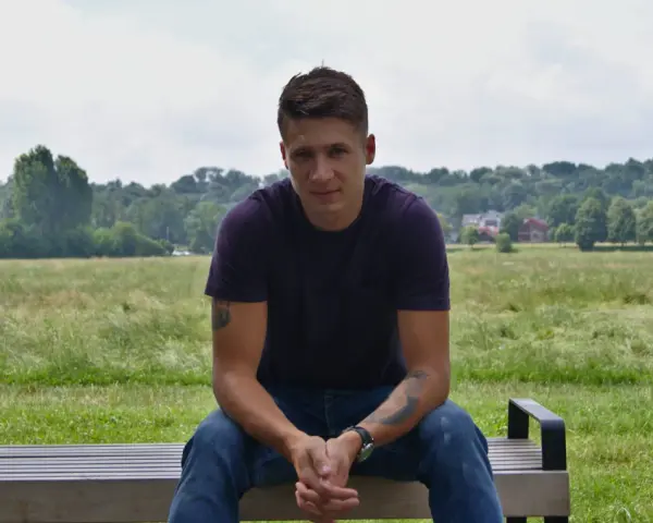 Bartłomiej Bis siedzi na ławce w parku, w tle zielone drzewa i niebo.