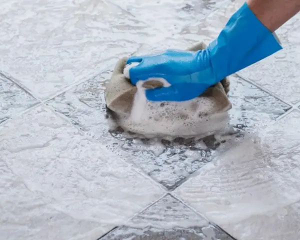 Mano con guanto blu che pulisce ceramica macchiata con spugna e schiuma.