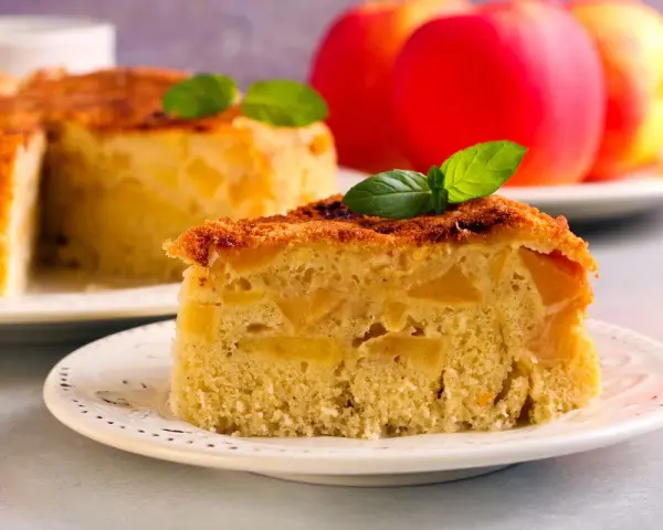 Una fetta di torta di mele vegana, soffice e ricca di mele, guarnita con menta. Sullo sfondo, una torta intera e mele rosse.
