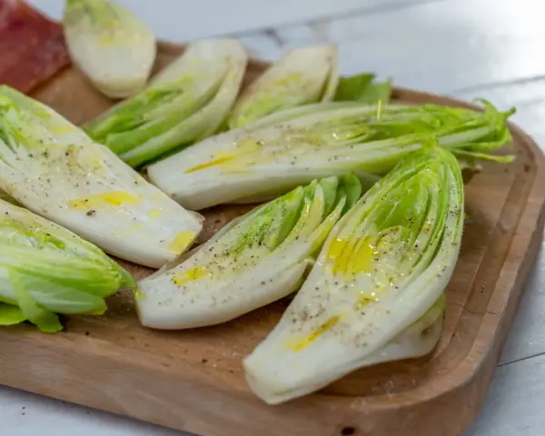 Insalata belga tagliata a metà, condita con olio e pepe, su un tagliere di legno.