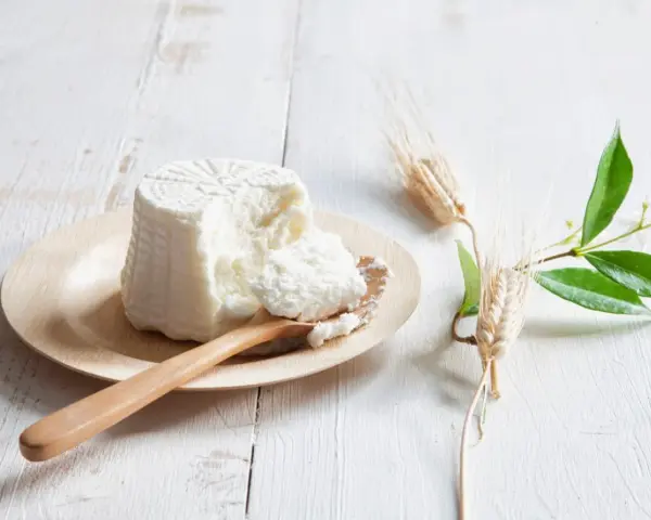 Świeża ricotta na drewnianej łyżce, obok kłosy zbóż i zielone gałązki. Idealna do dań, gdy szukasz czym zastąpić ricottę.