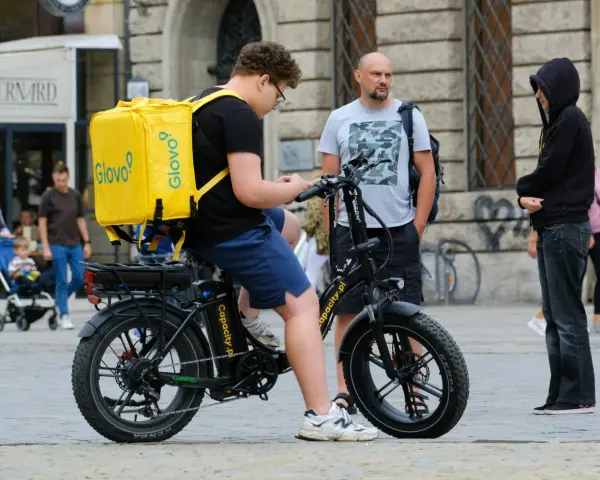 Młody kurier Glovo na rowerze elektrycznym sprawdza telefon. Obok stoi mężczyzna, a w tle widać ludzi i budynki.