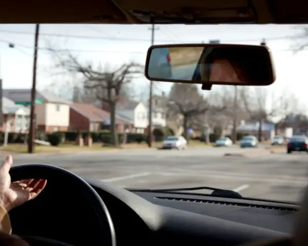 Mano sul volante, pronta per una manovra di svolta. Riflesso del volto nel retrovisore, case e alberi spogli fuori dal finestrino.