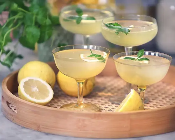 Fyra glas med en ljusgul limoncello drink, garnerade med mynta och citron. Citroner ligger på en träbricka.