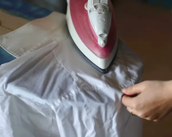 Różowe żelazko na desce do prasowania, obok biała koszula. Dłoń poprawia materiał, pokazując, jak prasować koszule.