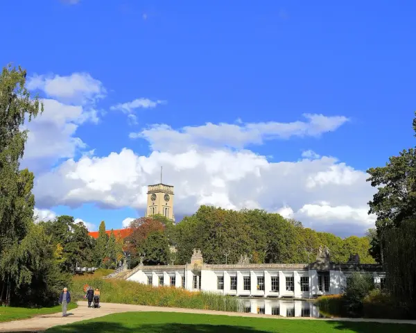 Ein sonniger Tag im **Rudolph Wilde Park**. Ein langer, weißer Pavillon spiegelt sich im Wasser, während ein Uhrenturm über Bäumen aufragt.