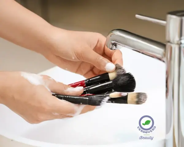 Mani che lavano pennelli da trucco insaponati sotto un rubinetto cromato. Impara come pulire i pennelli da trucco per una pelle sana.
