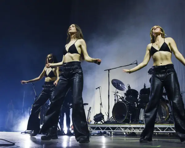 Drei Frauen in Lederhosen und BHs performen auf der Bühne, ein Schlagzeug im Hintergrund. Sie sind wie eine moderne Metal-Band.