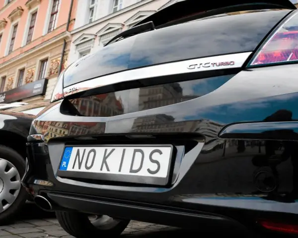 Czarne auto z indywidualnymi tablicami rejestracyjnymi "NO KIDS" na tle kamienicy.