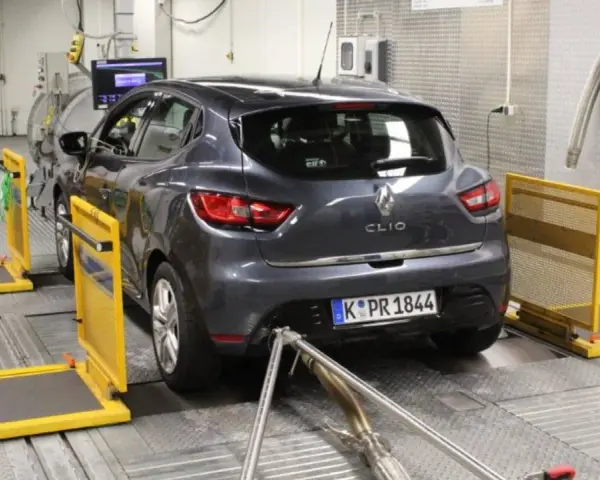 A dark grey Renault Clio is on a dynamometer in a testing facility, with exhaust pipes connected.