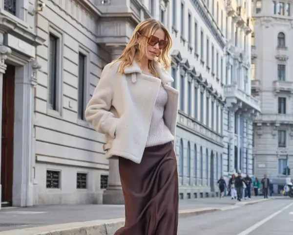 Donna con cappotto bianco in stile teddy, gonna di seta marrone e occhiali da sole, passeggia in città. Il **capispalla** è un elemento chiave per definire lo stile.