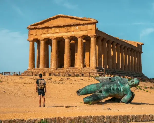 A lone traveler explores the Valley of the Temples in Sicily, standing before an ancient Greek temple and a fallen bronze statue.
