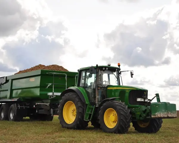 Zielony traktor John Deere z przyczepą wypełnioną ziemniakami na polu.