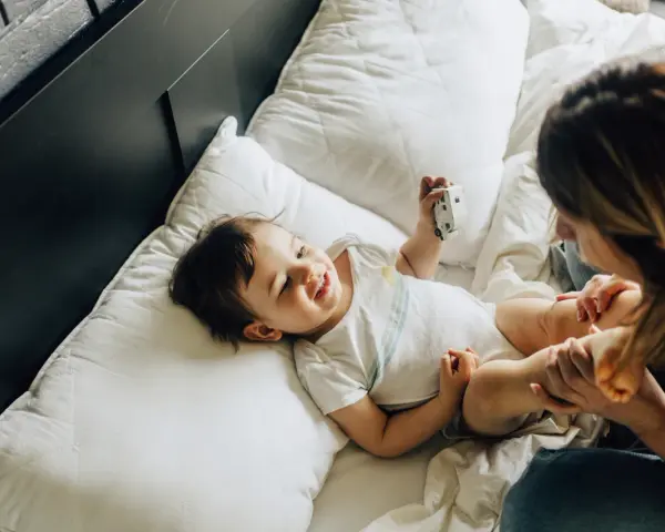 Uśmiechnięte dziecko bawi się zabawką na łóżku. Mama patrzy na malucha. Co to za mastka?