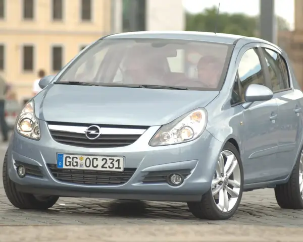 Jasnoniebieski Opel Corsa D z włączonymi żarówkami z przodu, zaparkowany na bruku.