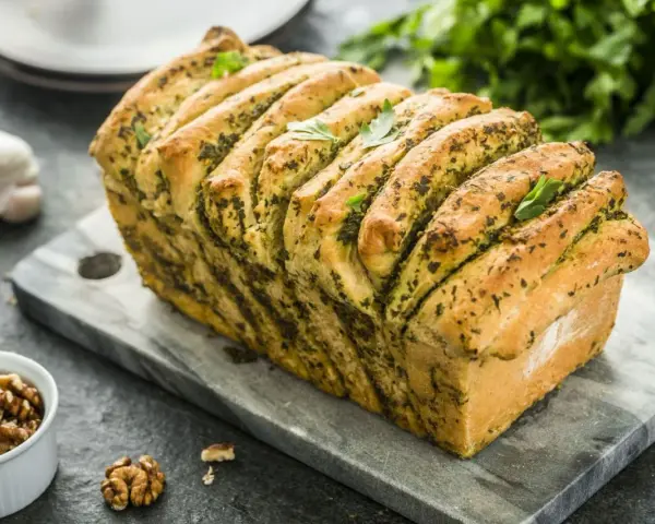 Pyszny chlebek rwany z pesto, udekorowany listkami bazylii, prezentuje się apetycznie na kamiennej desce. Obok czosnek i orzechy włoskie.