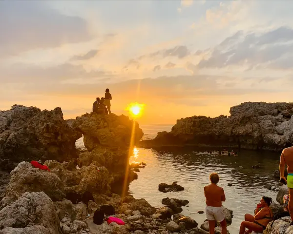 Tramonto dorato sulla piscina di Venere a Milazzo. Persone sedute sulle rocce e in acqua, godendosi la vista.