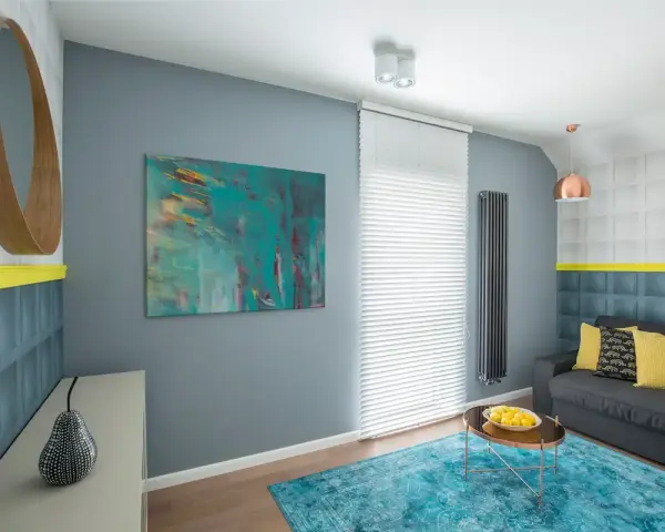 Sala de estar con sofá gris, alfombra turquesa y arte abstracto en tonos fríos. Un espejo redondo de madera y detalles en amarillo complementan el espacio.