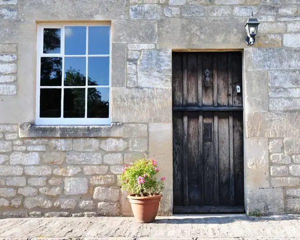 Facciate in pietra e mattoni grezzi con una porta in legno scuro e una finestra bianca. Un vaso di fiori rosa aggiunge un tocco di colore.