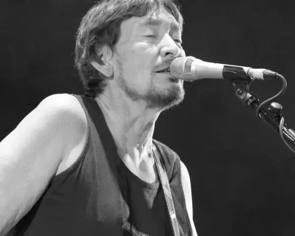 Chris Rea nie żyje. Muzyk śpiewa do mikrofonu na czarnym tle, w czarnej koszulce bez rękawów.