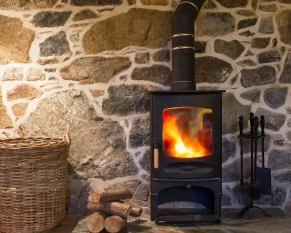 Stufa a pellet accesa su muro in pietra. Con cesto di vimini e attrezzi per camino. Potrei installare una stufa a pellet da solo.