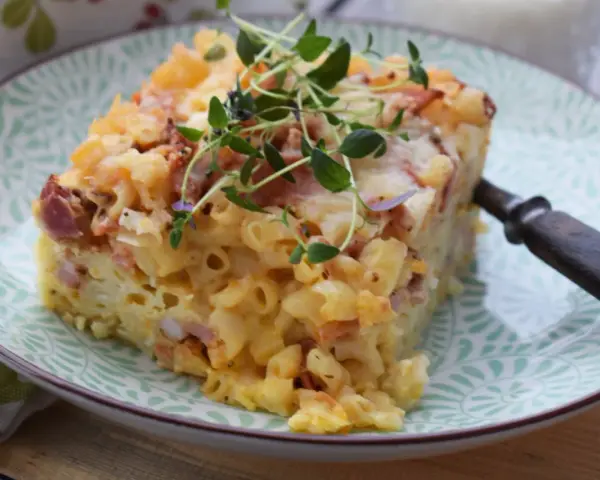 En bit makaronipudding med falukorv, toppad med färska örter, serveras på en grönmönstrad tallrik.