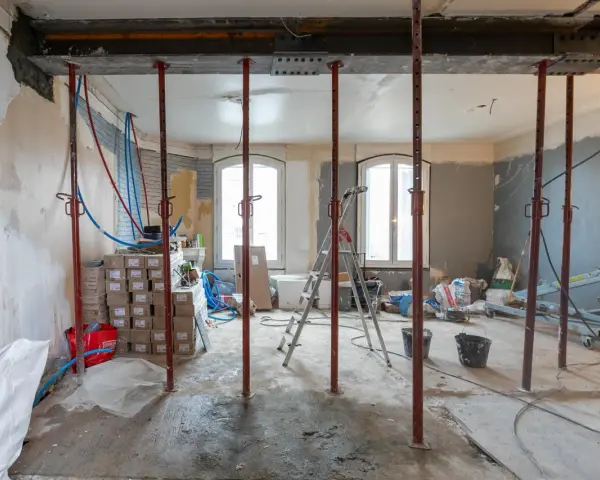 Chantier d'une maison phenix 1970 : poutres métalliques, étais, tuyaux et matériaux de construction dans une pièce en rénovation.