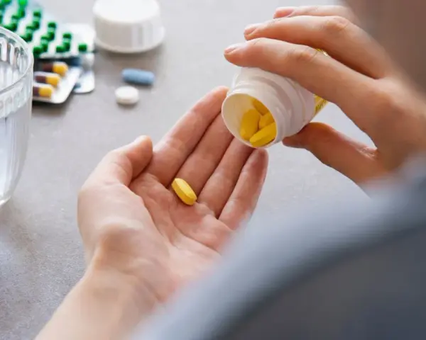 Dłoń wysypująca żółte tabletki z pojemnika na drugą dłoń, obok szklanka wody i opakowania leków.