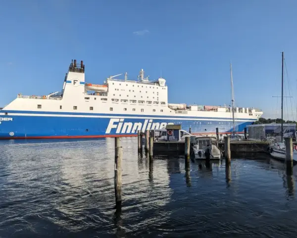 Duży prom Finnlines "Finntrader Malmö" cumuje w porcie, gotowy do rejsu. W tle widać żaglówki. Prom do Finlandii czeka na pasażerów.