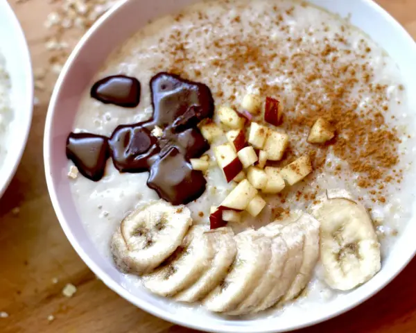 Un tazón de avena cremosa, perfecto para aprender como hacer porridge. Decorado con rodajas de plátano, trocitos de manzana, canela y un toque de chocolate derretido.