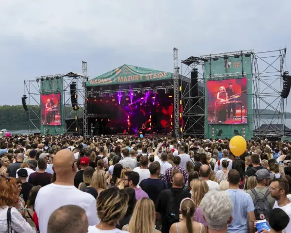 Tłum ludzi na koncercie pod gołym niebem. Na scenie gra zespół, a na ekranach wyświetlany jest green festival line up.