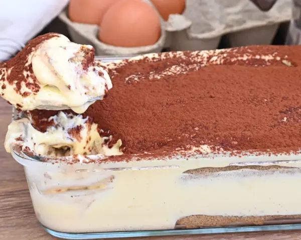 Un cucchiaio preleva una porzione cremosa di tiramisù, con 500 g di mascarpone, cosparso di cacao. Sullo sfondo, uova fresche.