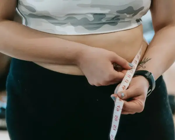 Kobieta mierzy swój duży brzuch centymetrem krawieckim, ubrana w sportowy top i czarne legginsy.