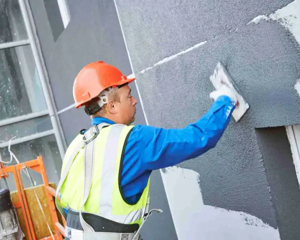 Obrero con casco naranja aplica revestimiento texturizado en fachada, parte de un sistema de aislante térmico exterior.
