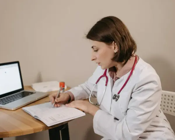Lekarka w białym kitlu z czerwonym stetoskopem siedzi przy biurku, pisząc w zeszycie. Obok leży laptop. Czy sor wystawia L4?